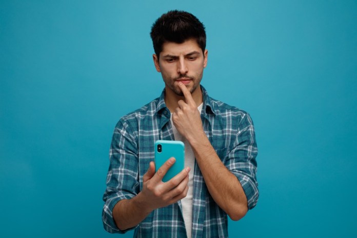 unsure young man holding and looking at mobile phone touching lip isolated on blue background