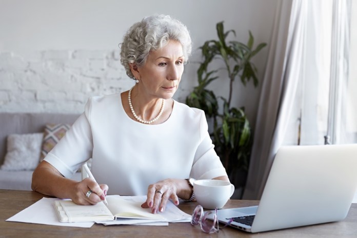 Aging, job, profession and electronic gadgets concept. Portrait of concentrated serious elderly retired businesswoman wearing pearl necklace looking at laptop screen and writing down in her diary