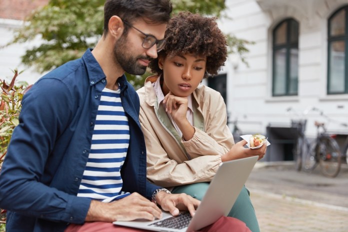 Outdoor shot of mixed race couple have attentive looks into laptop computer, use laptop computer, watch interesting video or movie, have snack, rest at bench against urban setting. Lifestyle concept