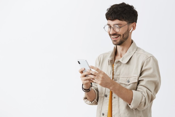Satisfied attractive caucasian male smartphone user in glasses and stylish jacket standing half-turned to left smiling joyfully with pleased look at smartphone screen wearing wireless earphones